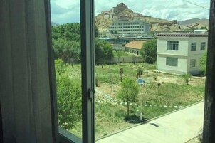 Vista a través de una ventana de un edificio con montañas al fondo, un área pavimentada con árboles y un panel solar.