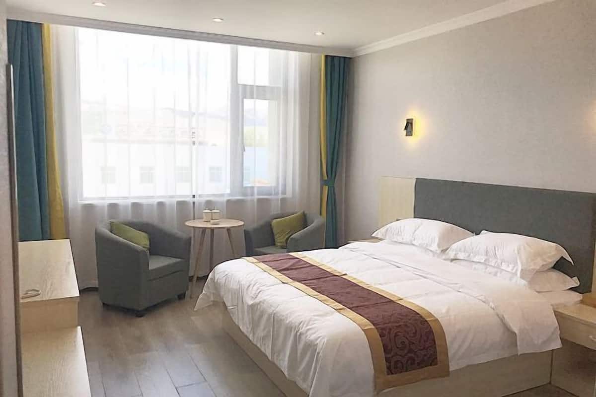 Een hotelkamer met een groot bed, een klein rond tafeltje, twee fauteuils en een raam met gordijnen.