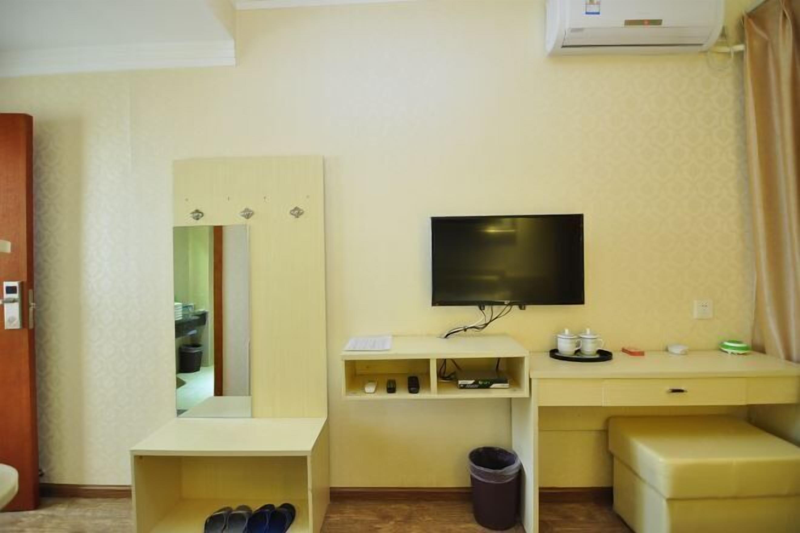 A hotel room with a TV, a desk, a mirror, a trash bin, and a shoe rack.