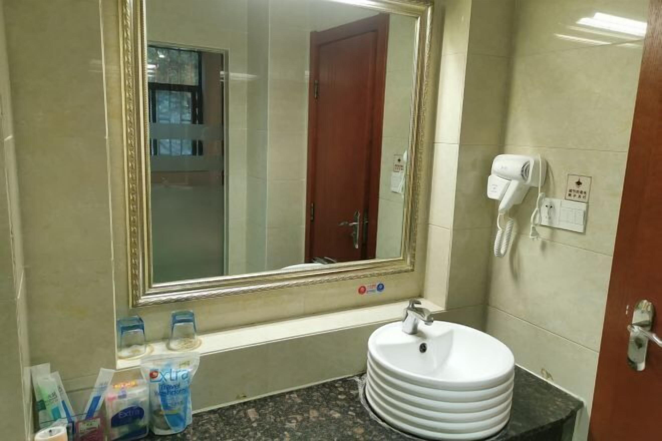 A bathroom with a large mirror, a sink with stacked plates, a hairdryer, and toiletries on the counter.