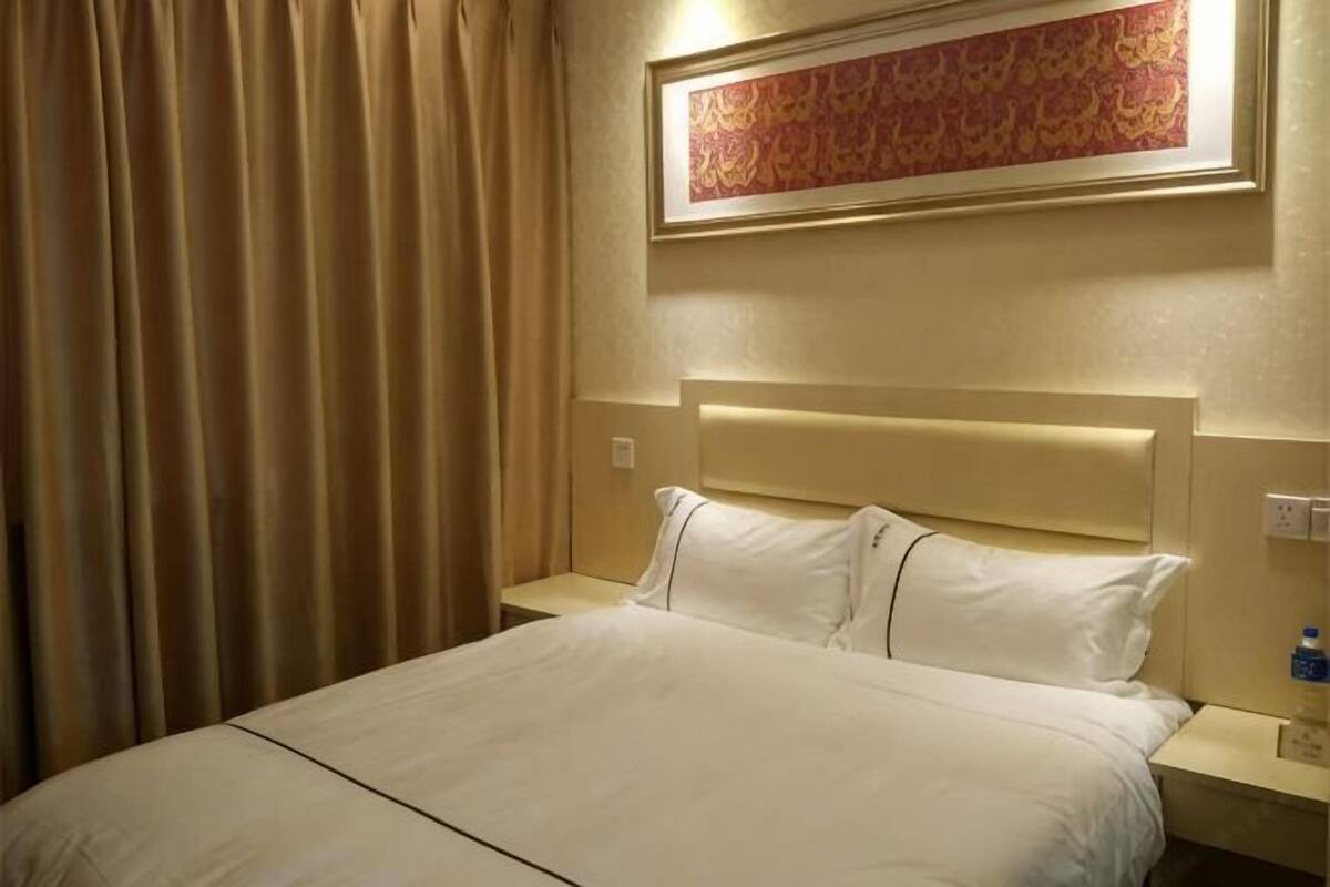A hotel room with a bed, curtains, a nightstand, a wall-mounted artwork, and a wall socket.