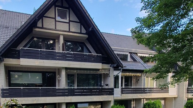 Ein modernes, zweistöckiges Haus mit Balkon, umgeben von einem gepflegten Rasen und kleinen Bäumen.