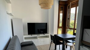 A modern living room with a sofa, a TV on a white stand, a dining table with chairs, and large windows offering a view of greenery. - Apartamentos Boutique Don Miguel (Noja)