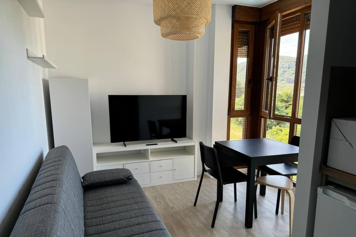 Un saló modern amb un sofà, una televisió sobre un suport blanc, una taula de dinar amb cadires i grans finestres que donen vista a vegetació.