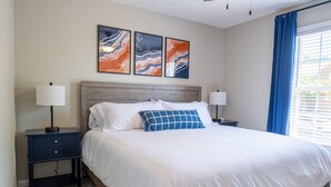 Un lit bien fait, avec une literie blanche et un coussin à carreaux bleus, flanqué de deux tables de chevet avec des lampes, dans une pièce dotée d’une tête de lit en bois et d’œuvres d’art abstraites accrochées au mur.
