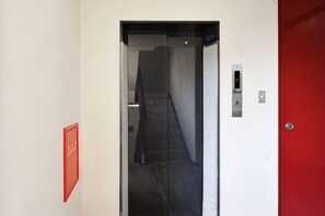A hallway with a red door, a staircase, and a wall-mounted elevator control panel. - Osaka House 701 | Near Namba, Direct to KIX! (Osaka)