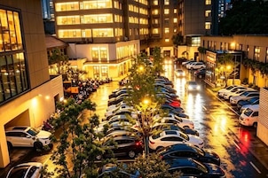 Um estacionamento com vários carros, um edifício com janelas iluminadas e uma árvore ao centro.