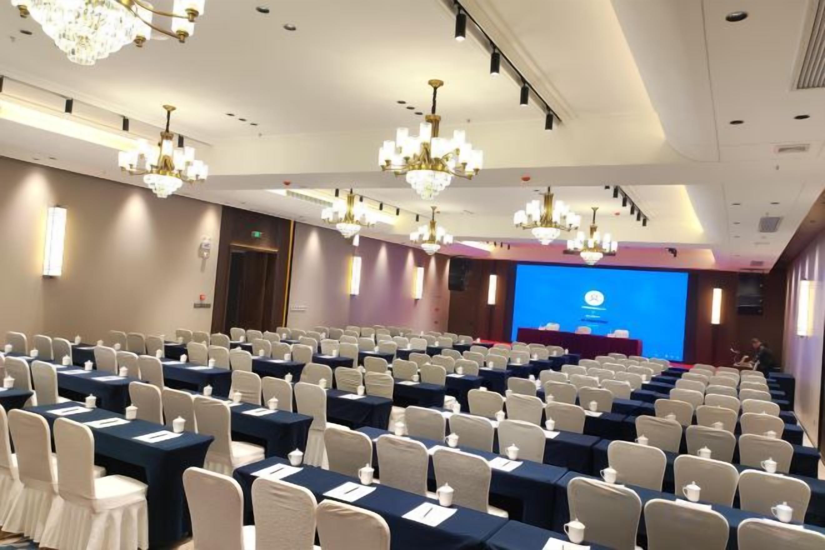 Een congreszaal met rijen tafels en stoelen, een podium en kroonluchters.