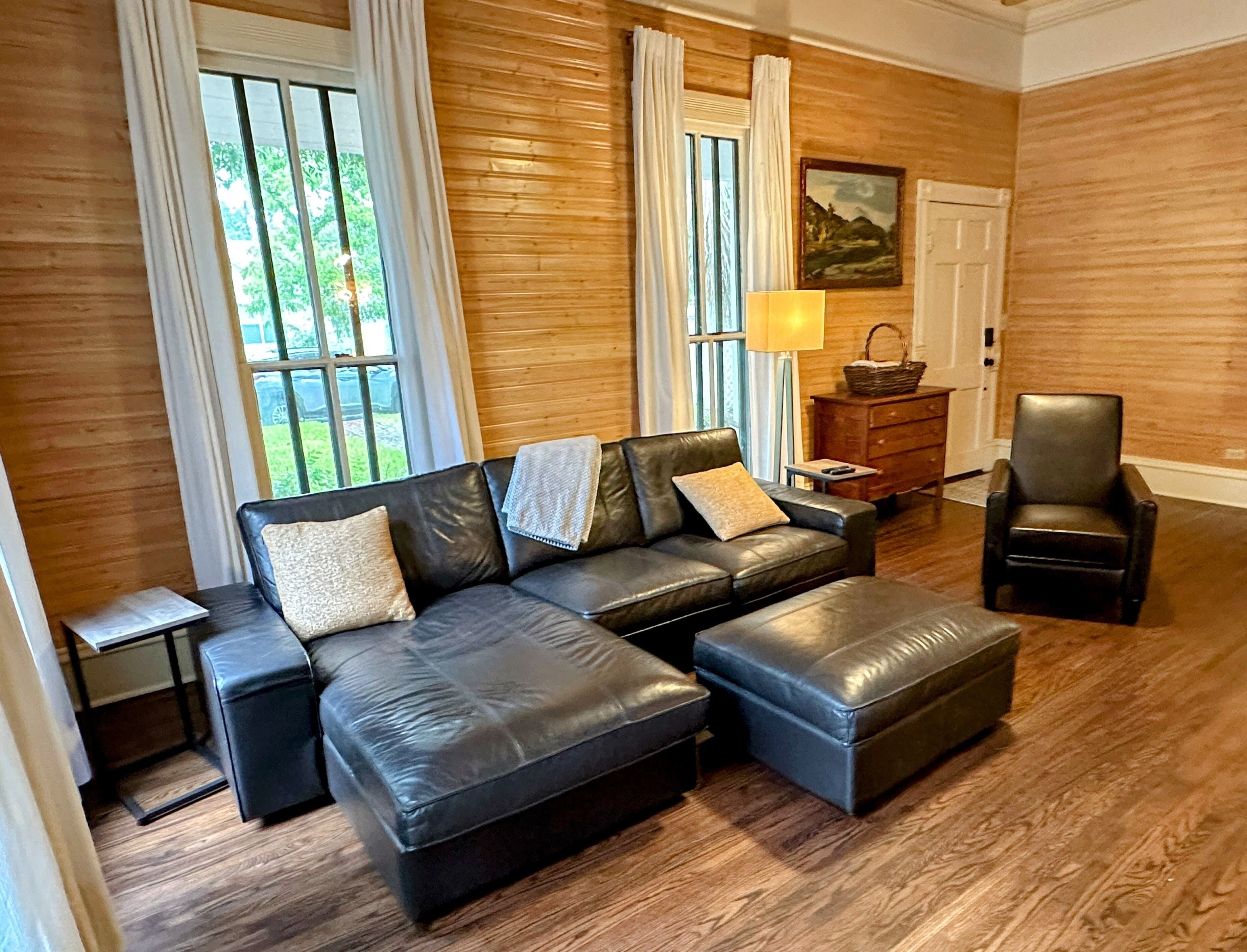 Una sala de estar moderna con un sofá seccional de cuero, un reposapiés a juego, piso de madera y ventanas grandes con cortinas.
