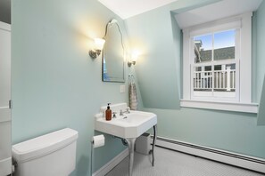 Un baño con inodoro y lavamanos blancos, un espejo con apliques de pared y una ventana con vista a una casa.