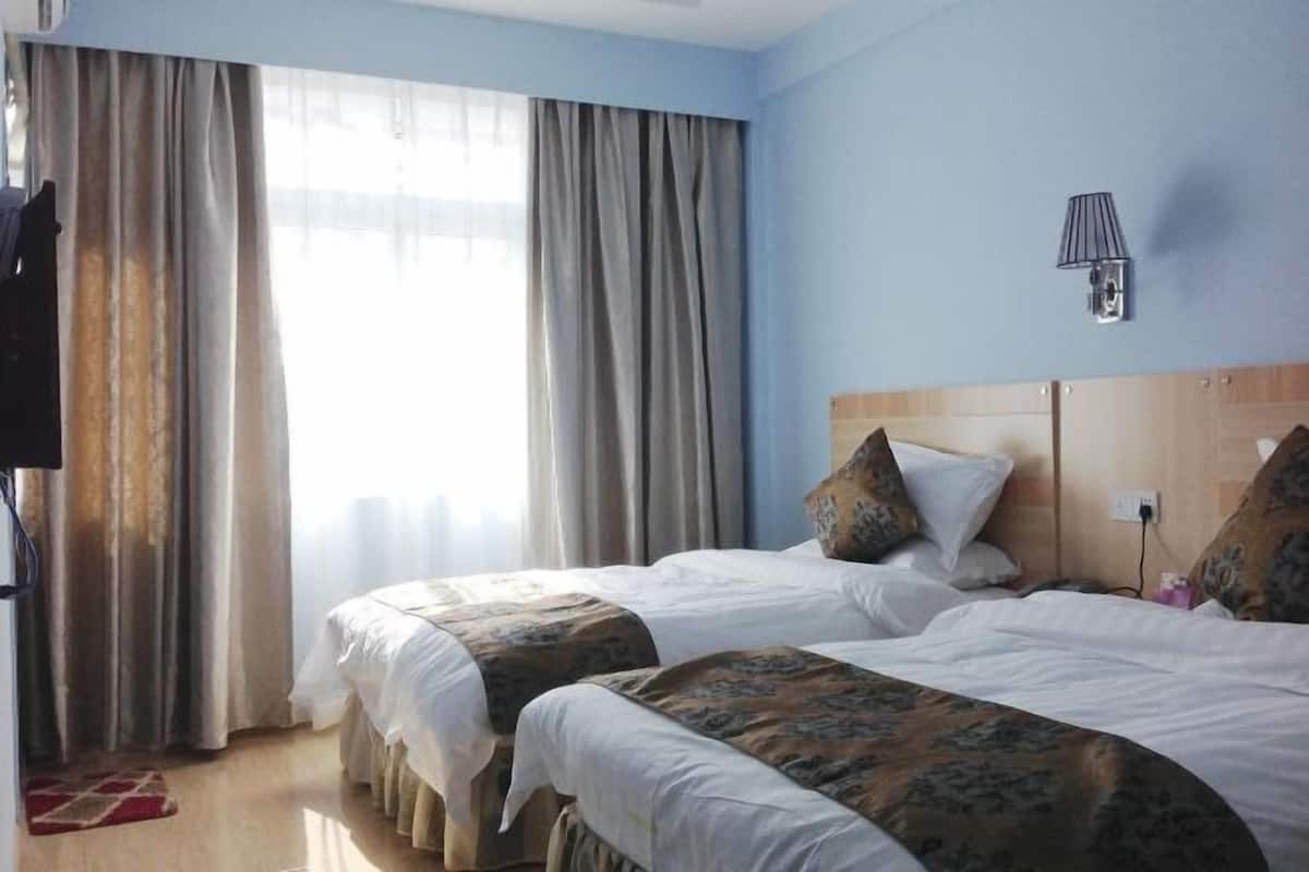 A hotel room with two beds, a wooden headboard, a wall-mounted lamp, and a window with curtains.
