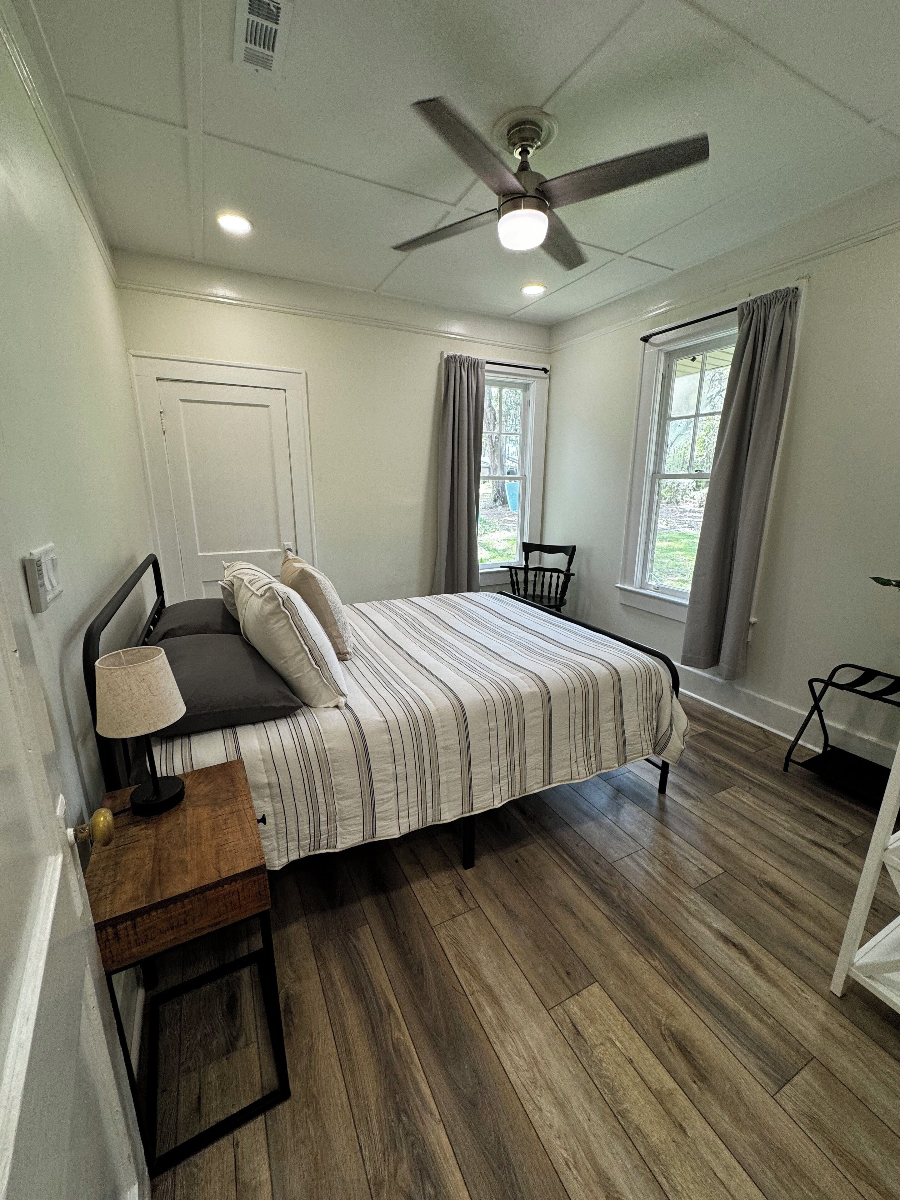 Un dormitorio con cama, un escritorio con lámpara, ventilador de techo y dos ventanas con cortinas.