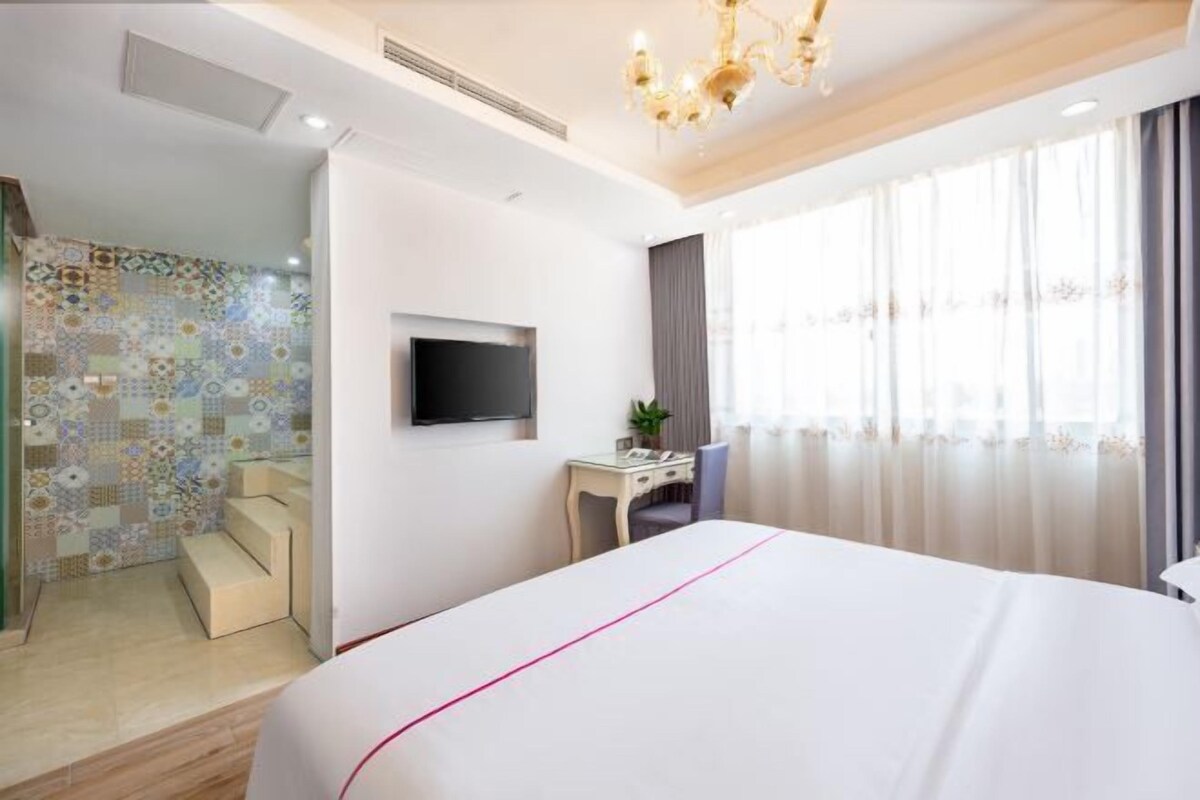 A hotel room with a large bed, a desk, a chair, a TV, and a shower area.