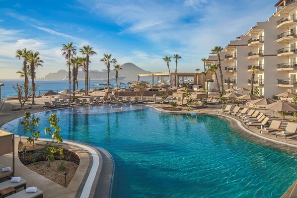 A resort pool with lounge chairs, palm trees, and a view of the ocean and mountains.