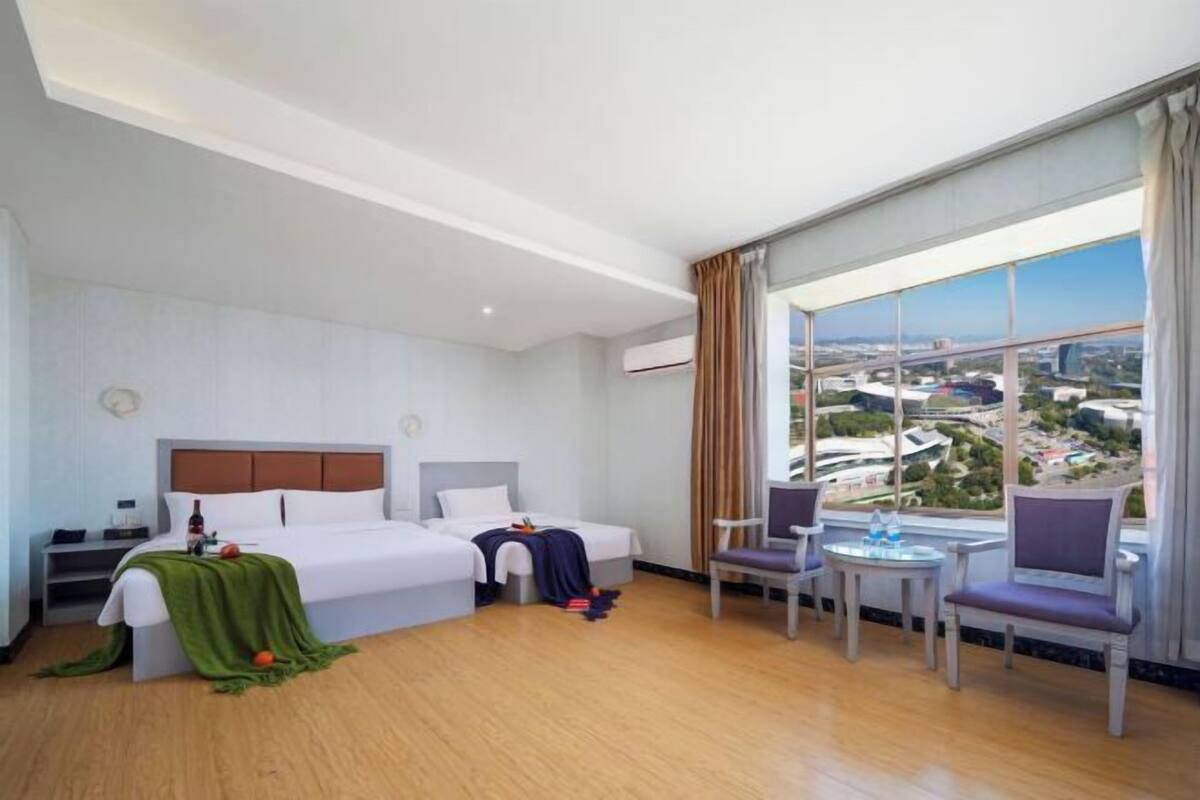 A hotel room with a large window, a bed, a chair, a small table, and a nightstand.