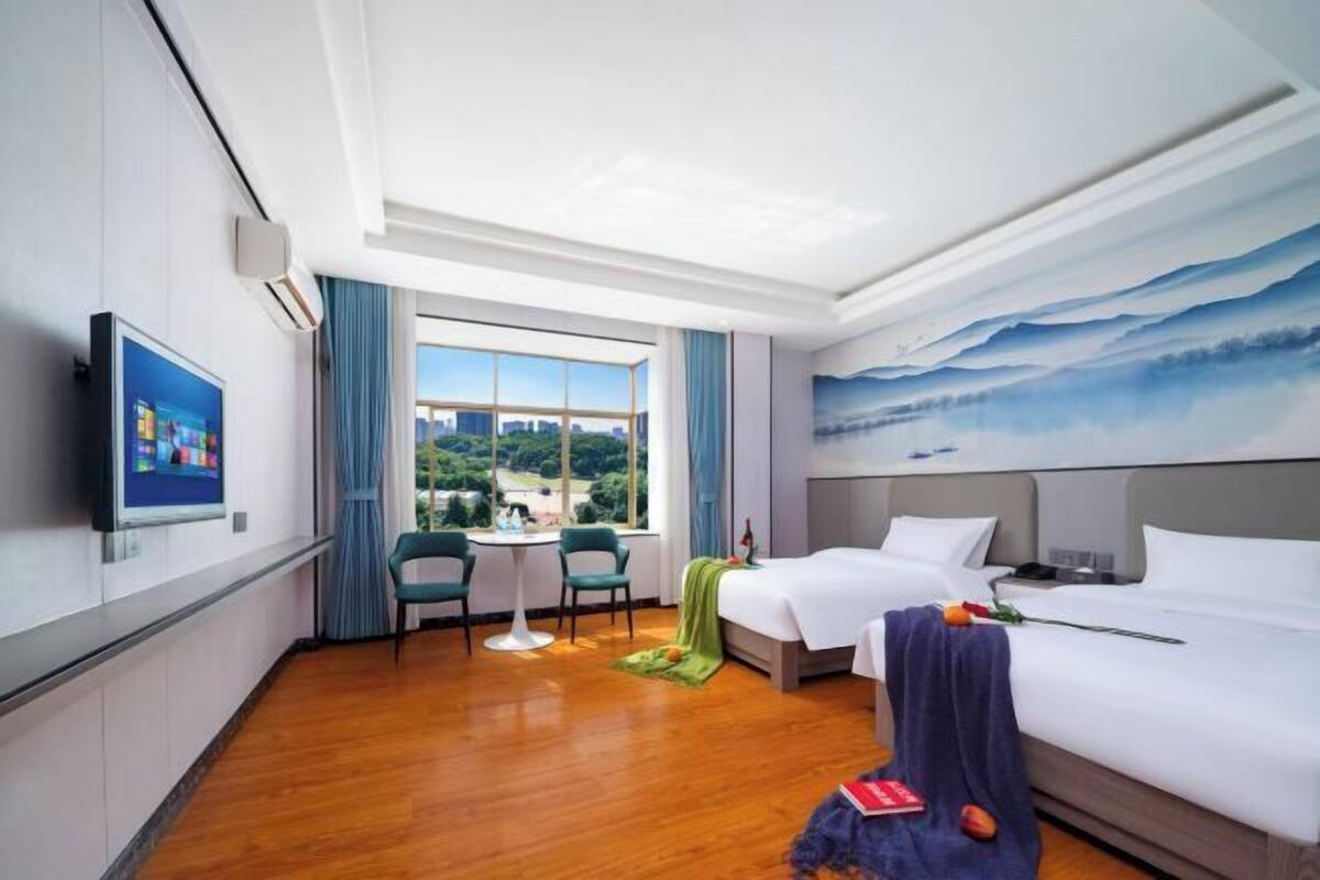 A hotel room with two beds, a TV, a window with a city view, and a painting on the wall.