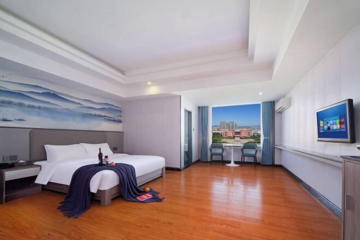 A hotel room with a bed, a nightstand, a TV, and a view of a cityscape through a window.