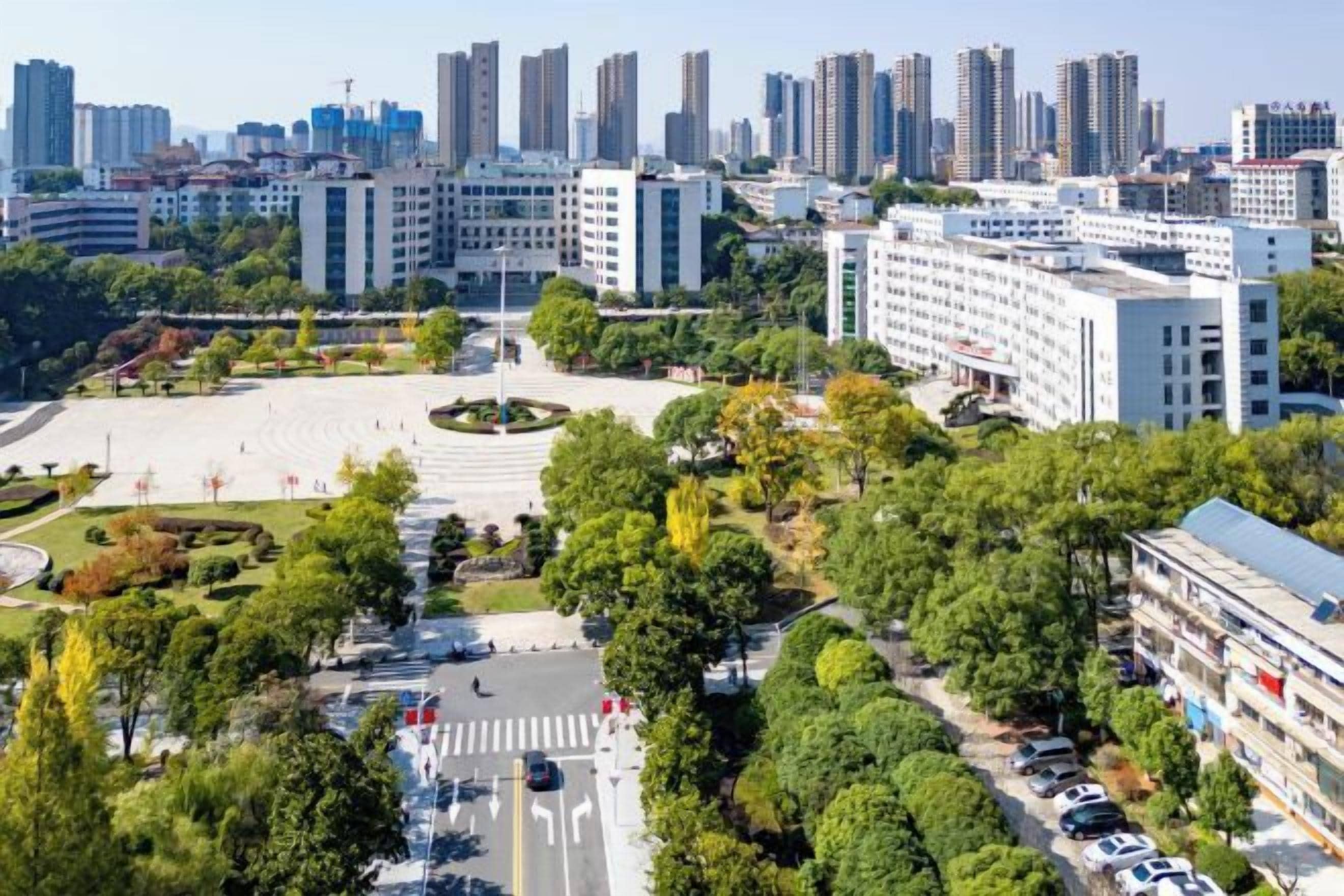 A cityscape with modern buildings, a central open plaza, and surrounding greenery.