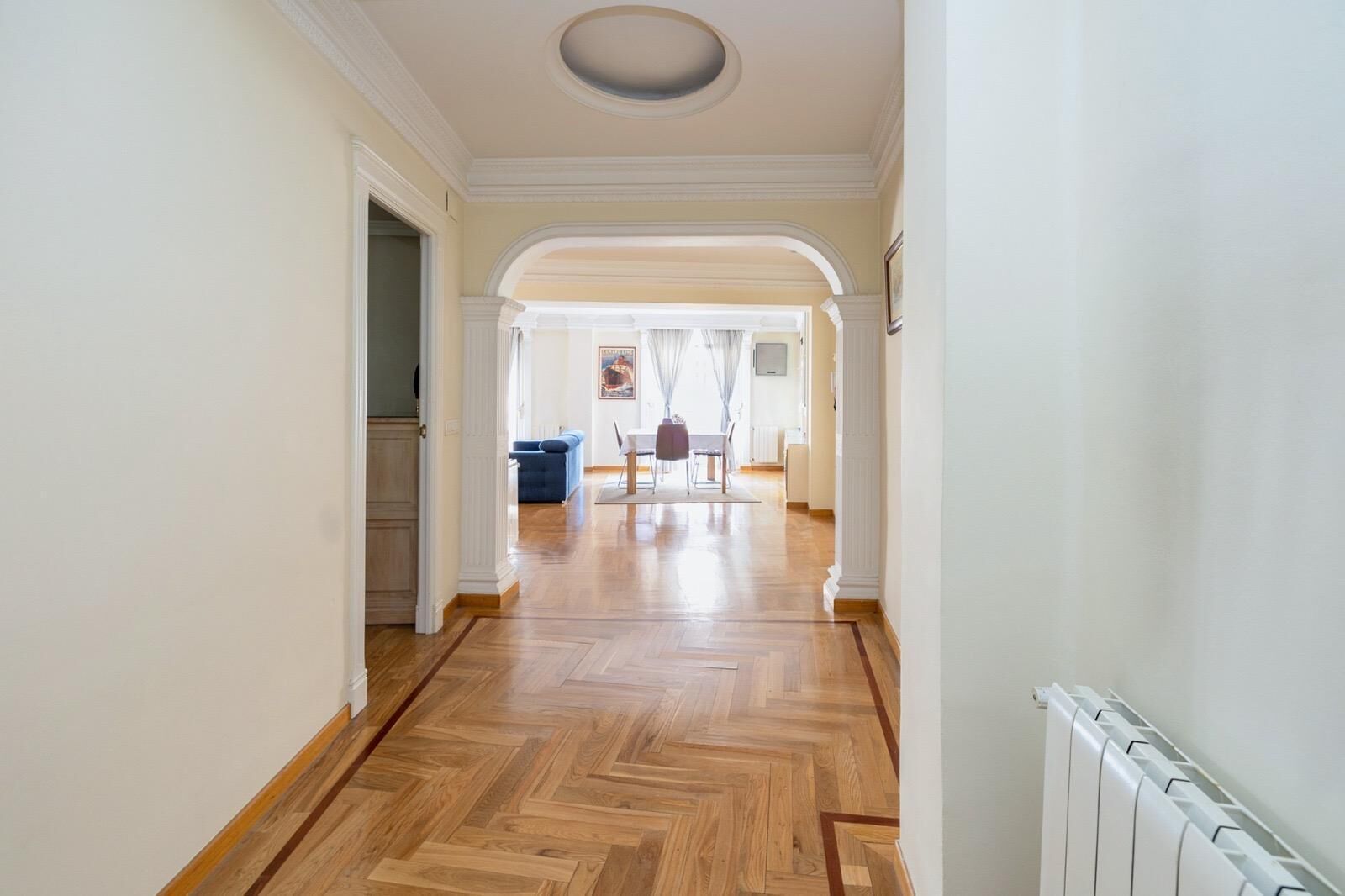 Un couloir au sol en bois et aux murs blancs, qui mène à un coin repas avec un canapé bleu et une table.