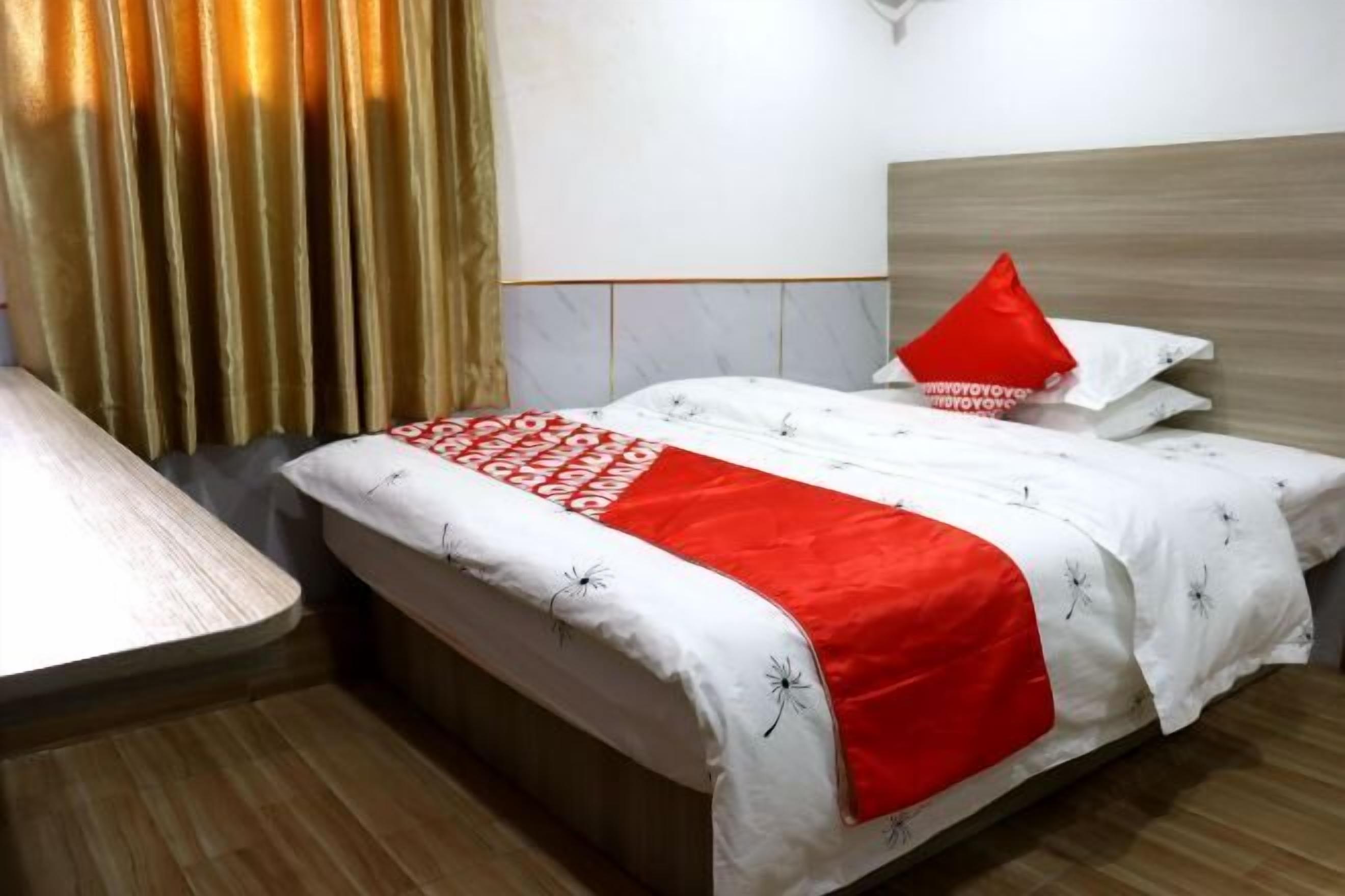 A hotel room with a bed, a bedside table, a red pillow, and a curtain.
