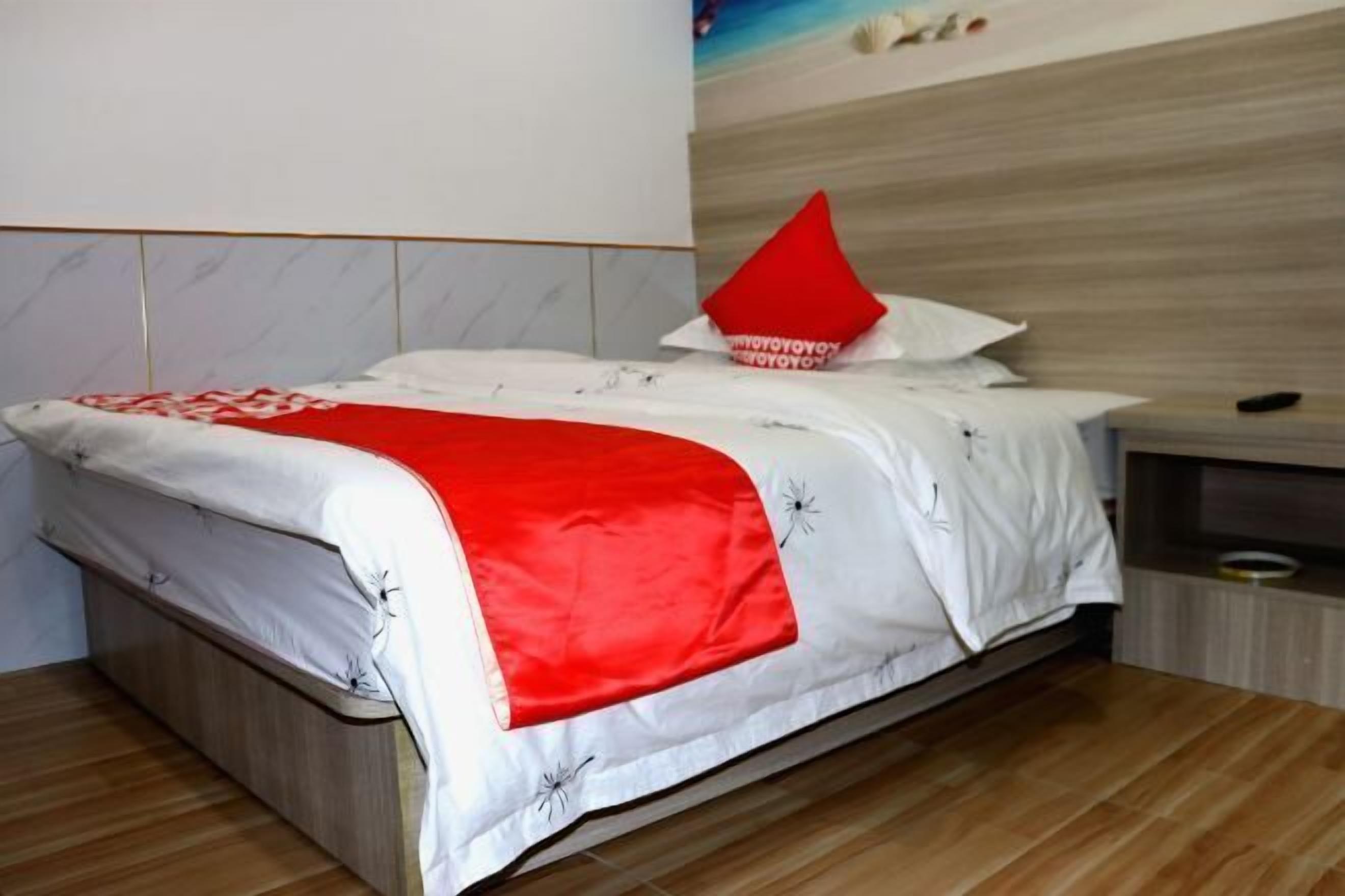 Una cama bien hecha con una colcha roja y blanca, un cabecero de madera y una mesita de noche con un control remoto.