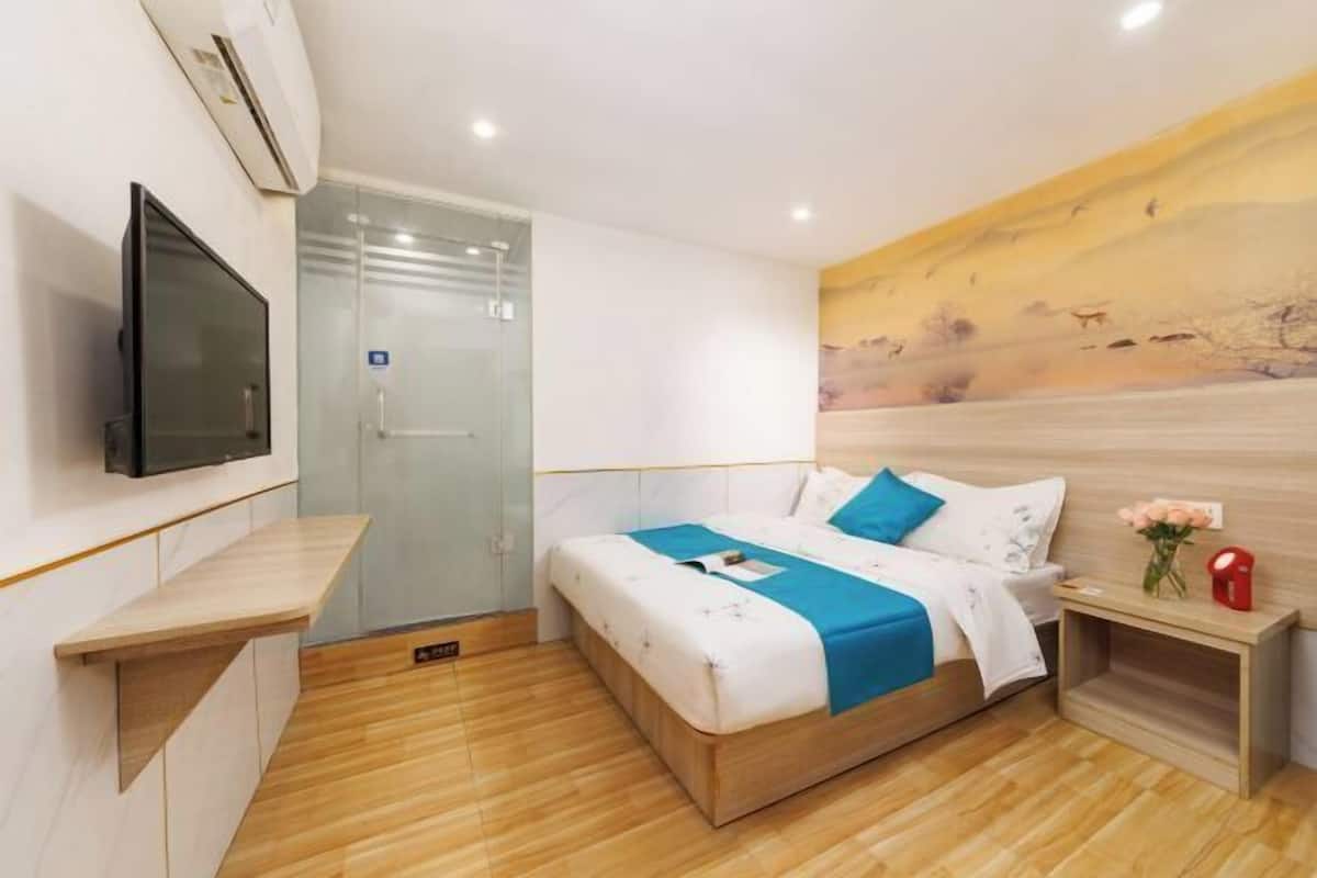 A modern hotel room with a bed, a TV, a glass shower enclosure, and a painting on the wall.