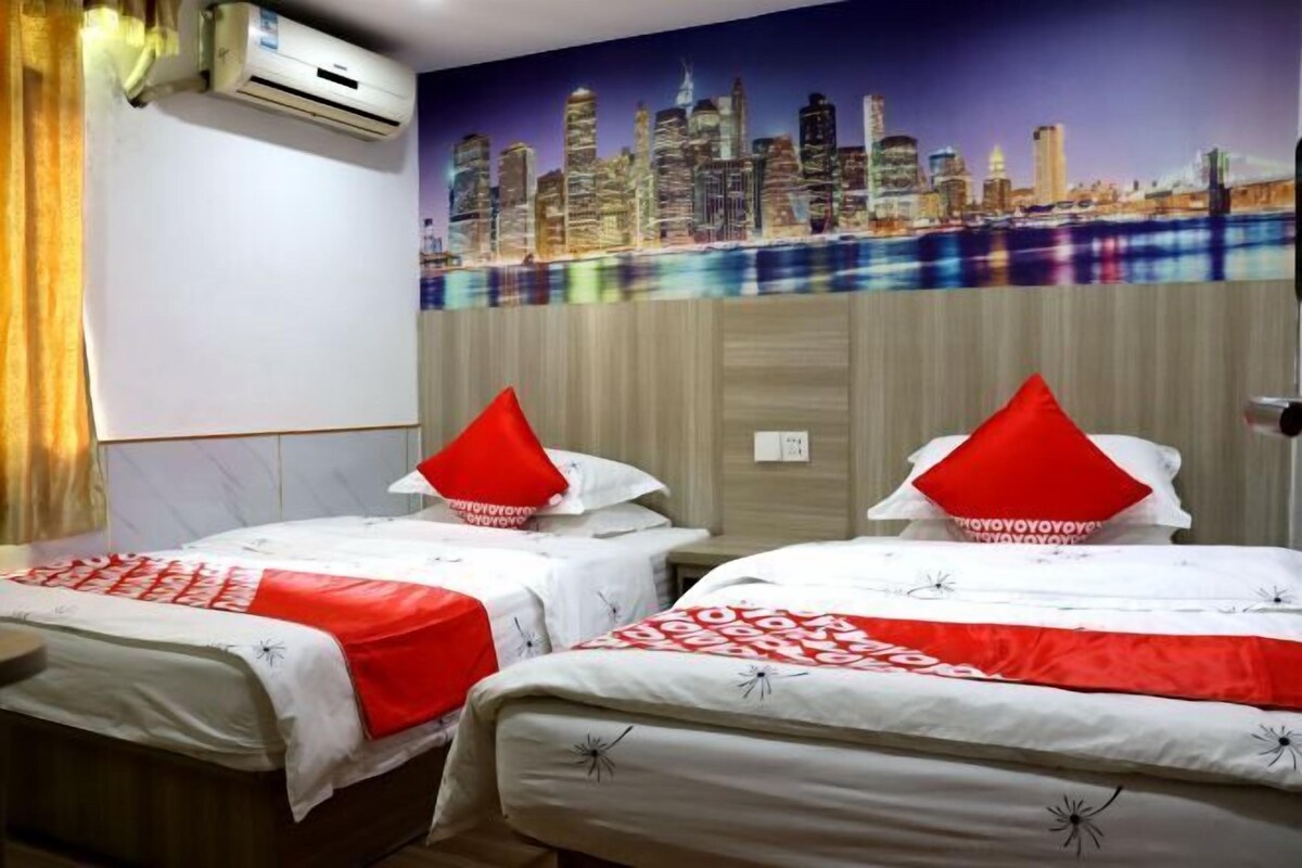 A hotel room with two beds, each with a red pillow, a cityscape mural on the wall, and an air conditioning unit.