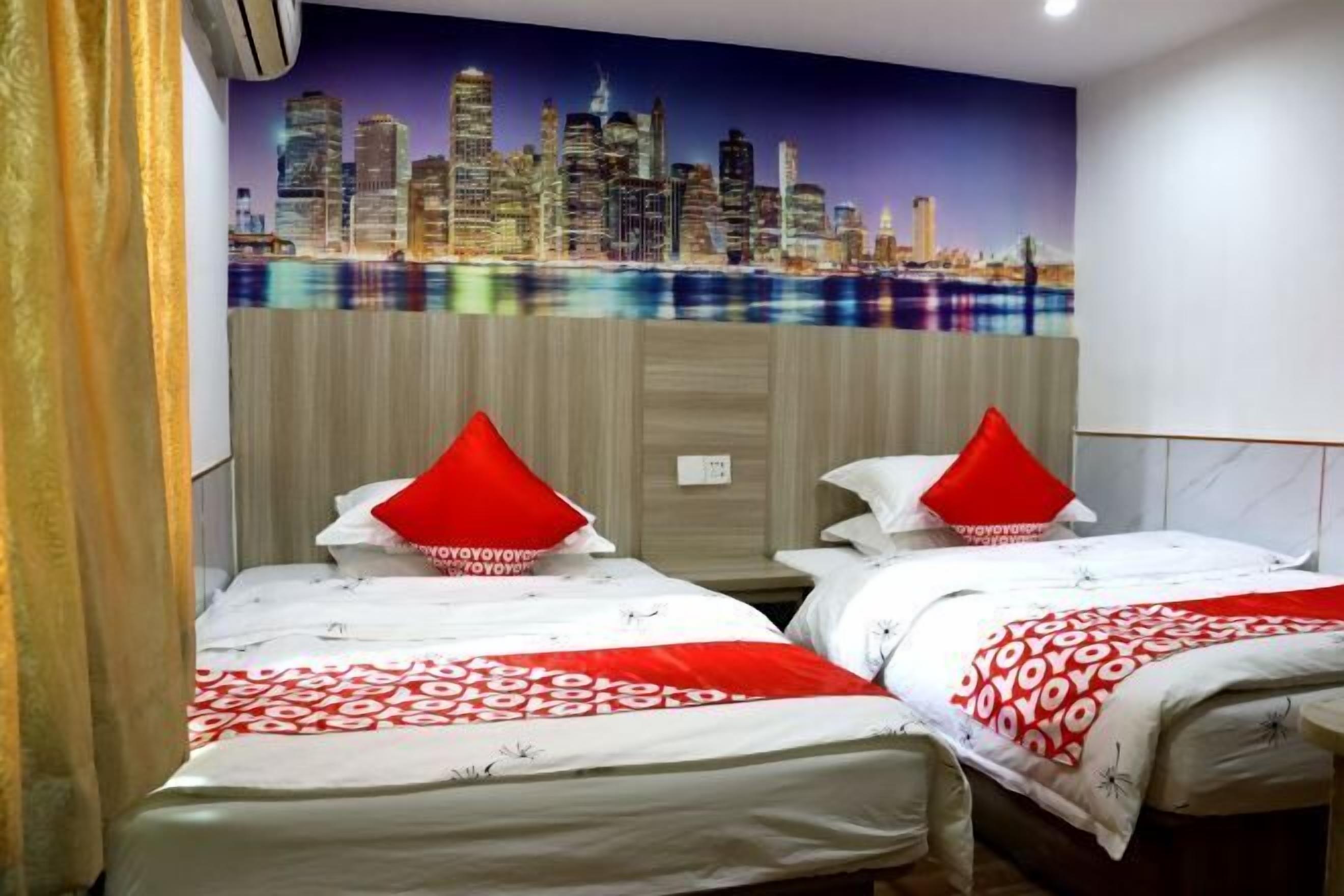 A hotel room with two beds, each with a red pillow and a patterned bedspread. The room features a cityscape mural on the wall and a wooden headboard.