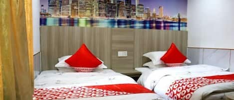 A hotel room with two beds, each with a red pillow and a patterned bedspread. The room features a cityscape mural on the wall and a wooden headboard.