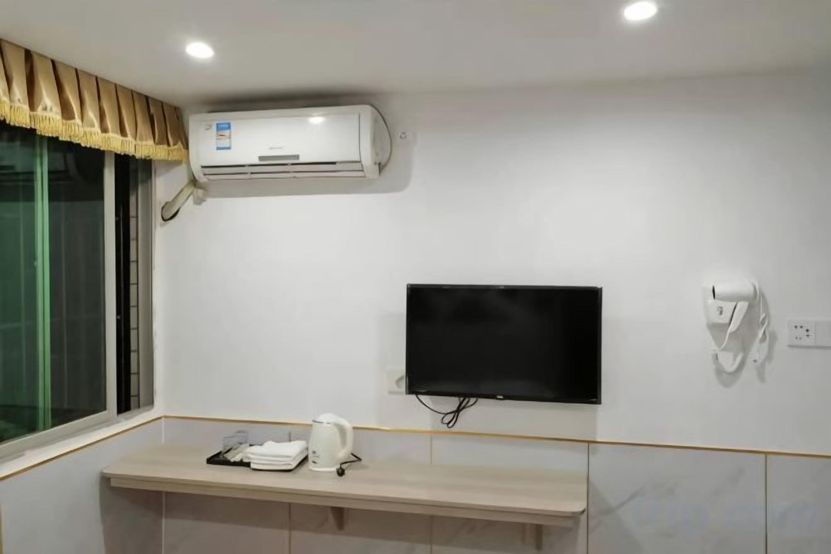 A hotel room with a mounted air conditioner, a flat-screen TV, a towel rack, and a small table with a kettle.