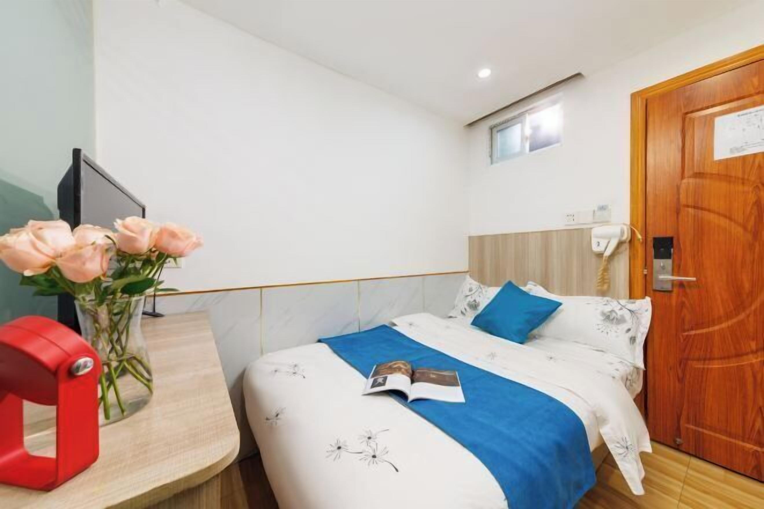 Une chambre d’hôtel avec un lit, une table de chevet ornée d’un vase de fleurs, un objet rouge posé sur la table et une porte en bois.
