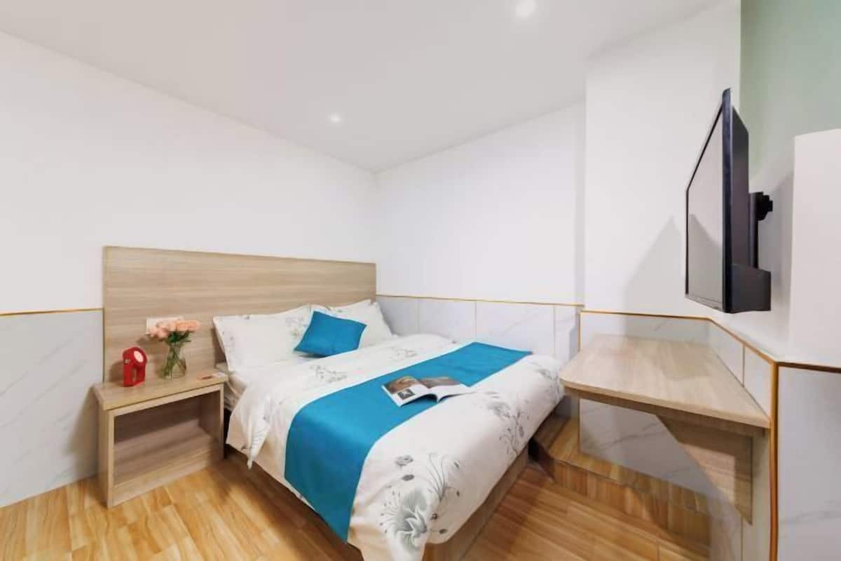 A modern hotel room with a wooden headboard, a bed with a blue bedspread, a bedside table with a red object, and a TV mounted on the wall.