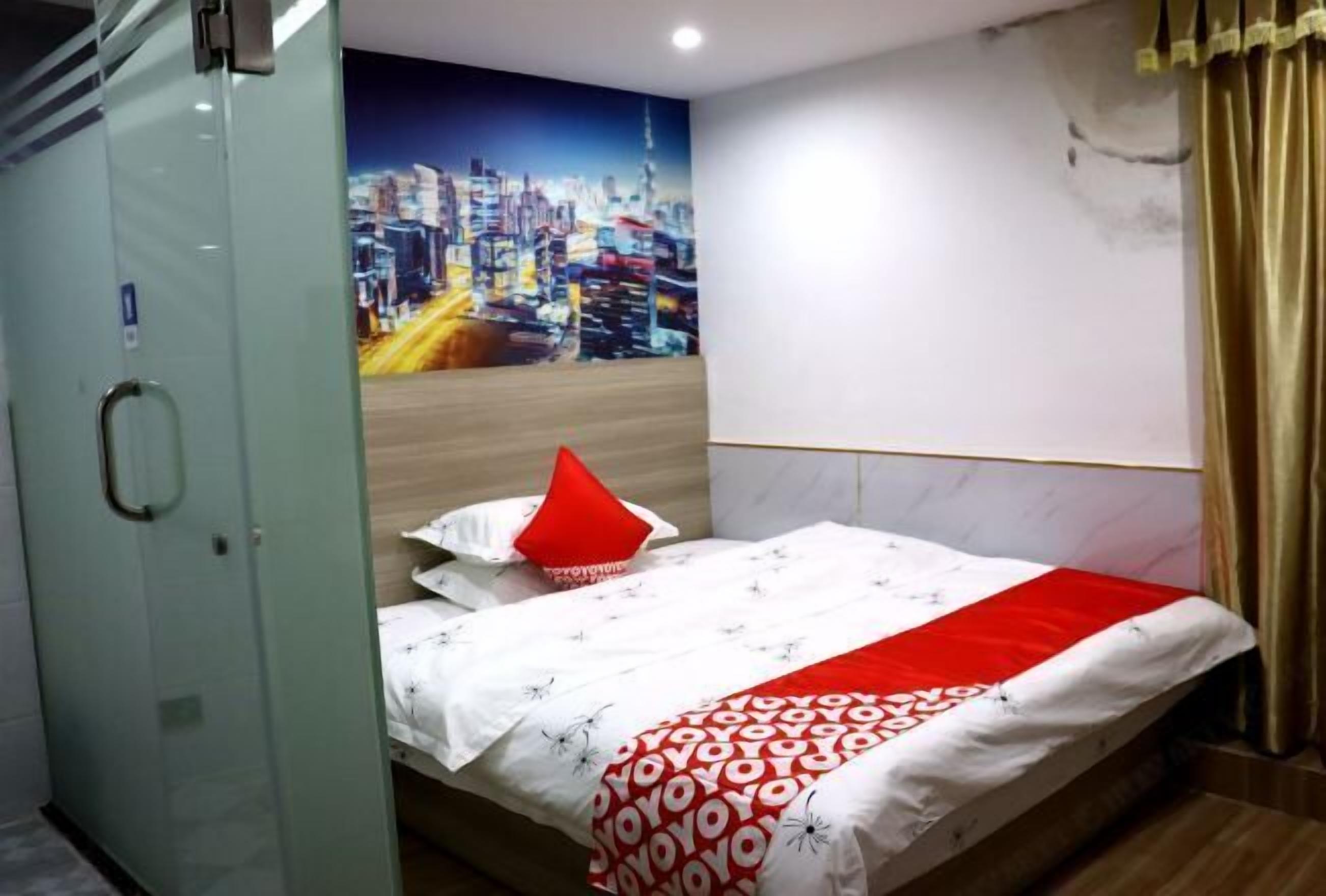 A hotel room with a bed, a cityscape mural, a red pillow, and a glass door.
