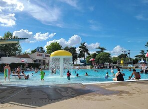A public swimming pool with multiple water features, including a central fountain, surrounded by lounge chairs and umbrellas. - Location! w/King Beds+Hot Tub+GameRoom+Theater+Gym+Fire Pit (Prairie Village)