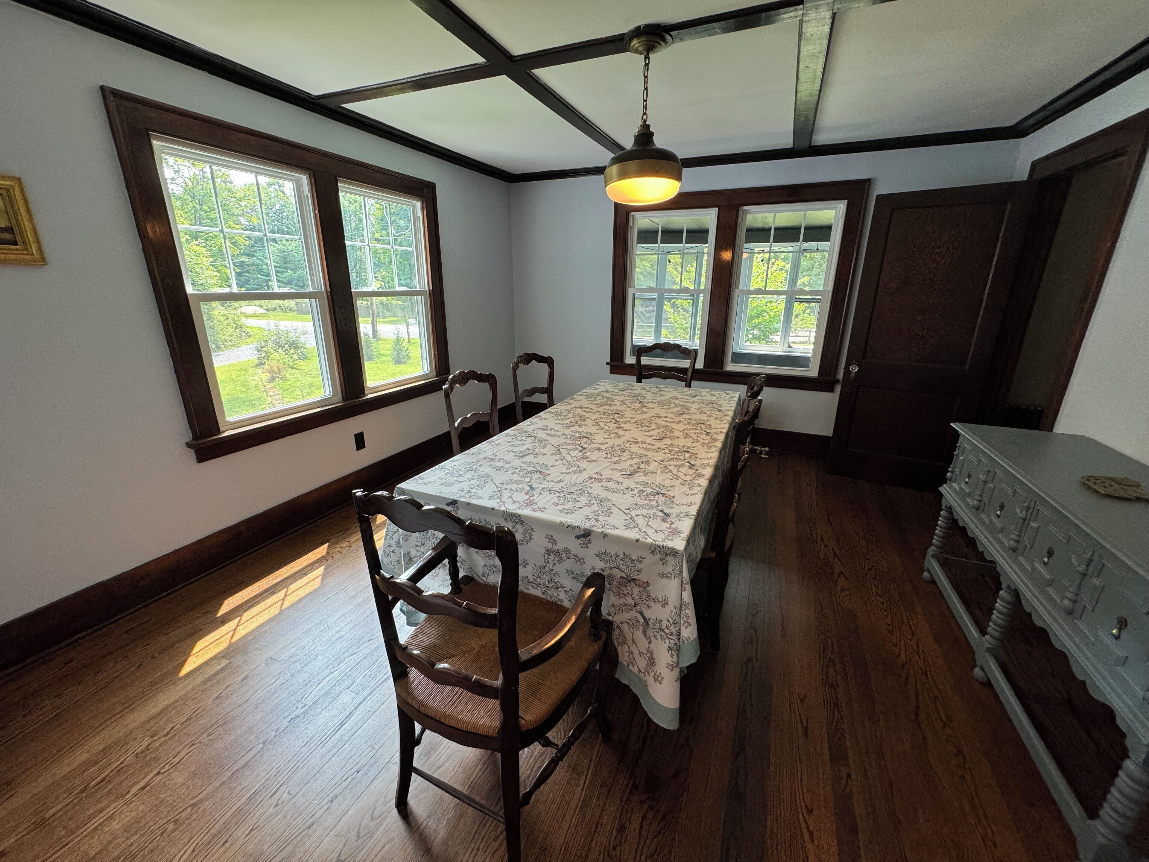 Une salle à manger avec une grande table, deux chaises et un lustre. Deux fenêtres donnent sur un paysage verdoyant.