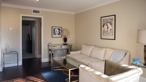 A living room with a beige sofa, a glass coffee table, and a lamp. There is a framed picture on the wall and a small table with a lamp.