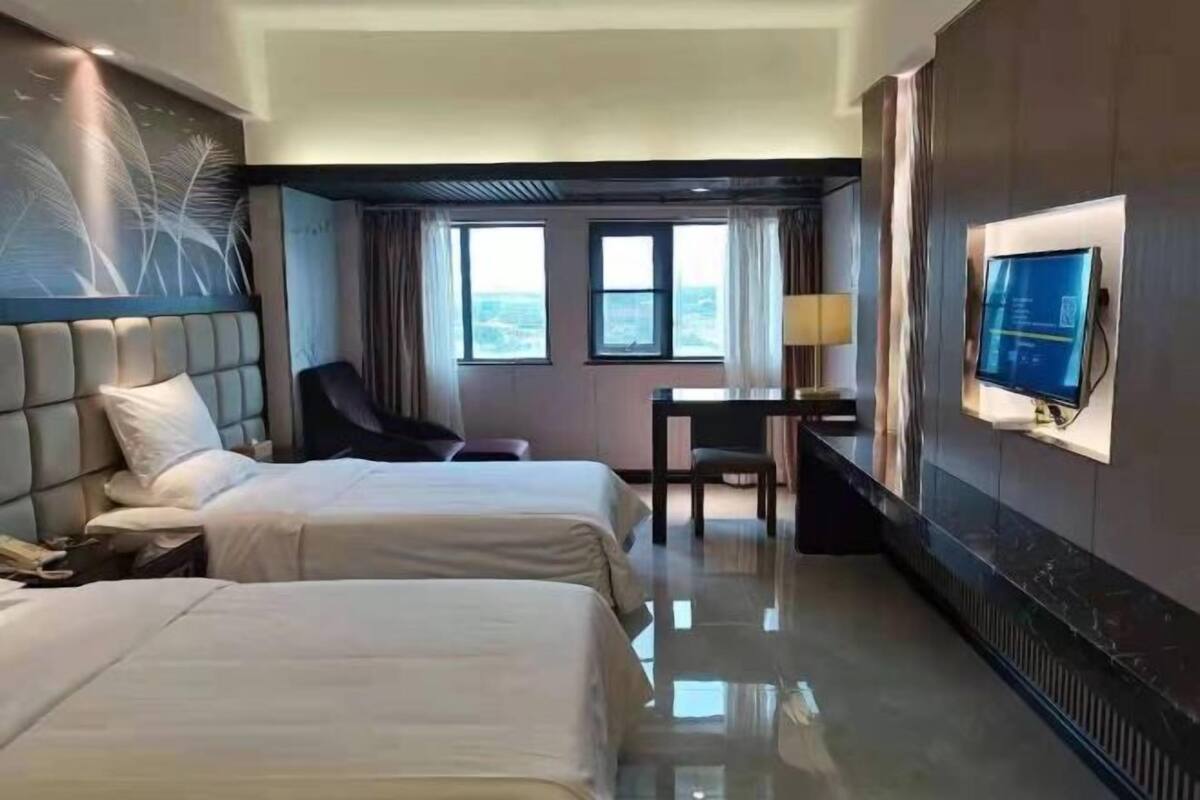 A hotel room with two beds, a desk, a chair, a TV, and large windows.