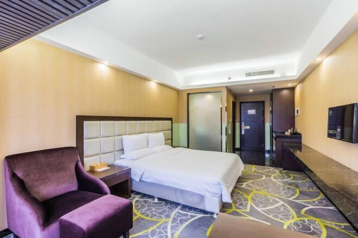 A hotel room with a bed, a purple armchair, a nightstand, and a bathroom door.