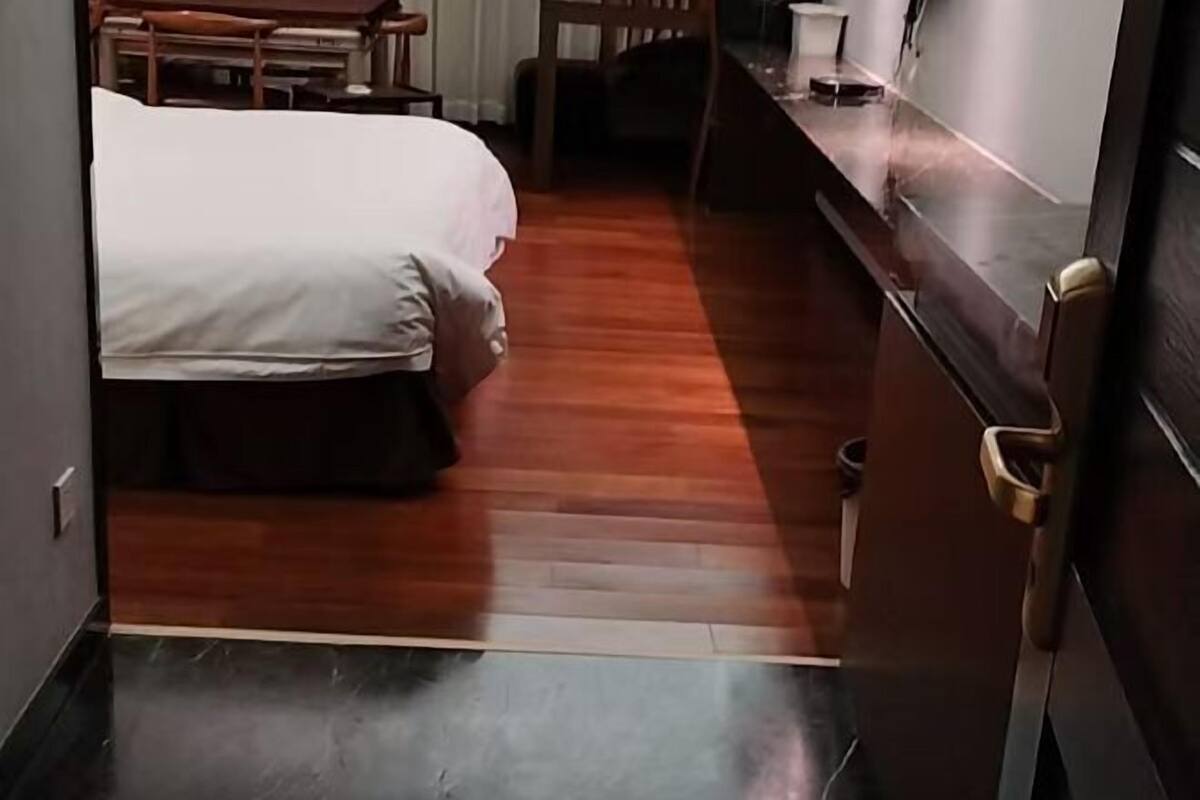 A hotel room with a wooden floor, a bed with a white bedspread, a wooden nightstand, and a door with a brass handle.