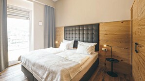 A modern bedroom with a large bed, a wooden headboard, a bedside table, and a view of the city through the window. - Apartment fuer 2 Personen (Brixen im Thale)