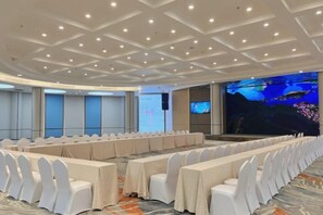 Un salón de conferencias con filas de sillas blancas, una pantalla grande y un escenario.