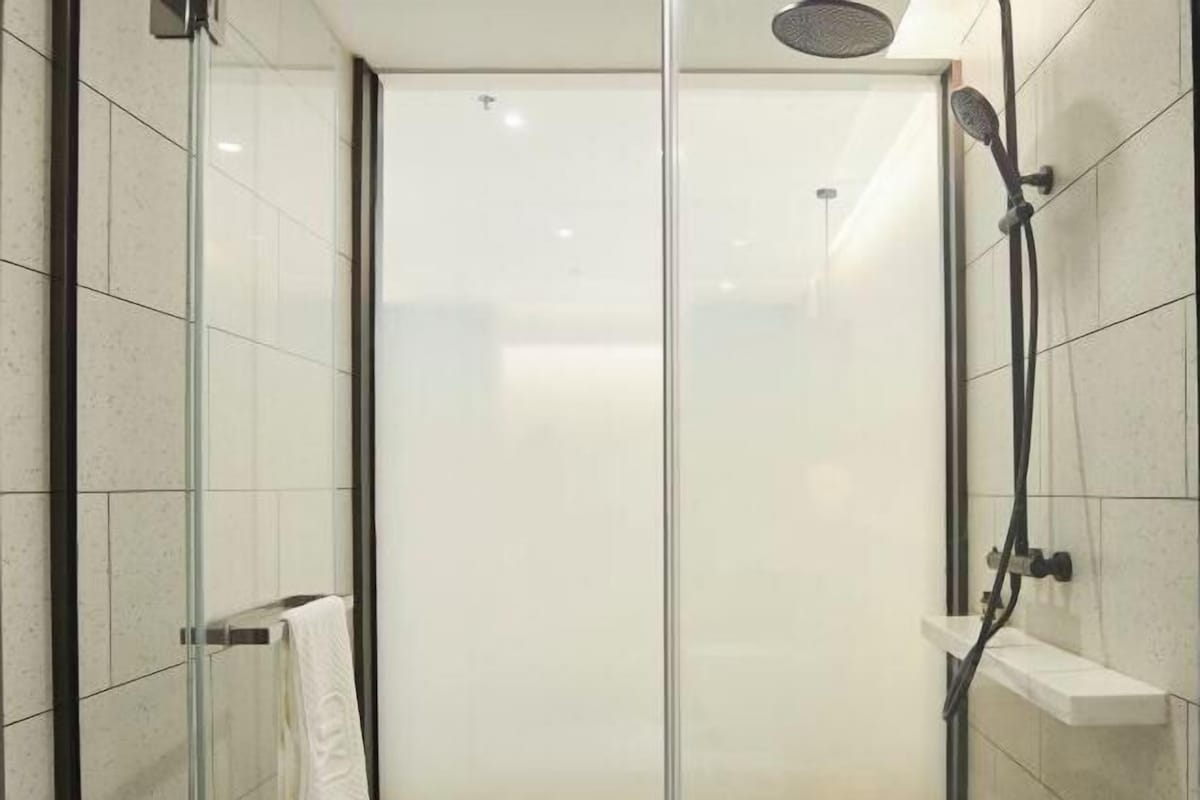 Cabina de ducha con paredes de vidrio, una ducha de mano, un perchero y un estante pequeño.