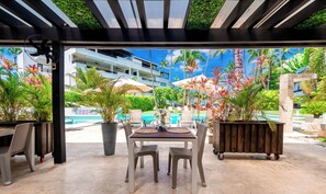 An outdoor patio with a table and chairs, surrounded by potted plants and umbrellas, overlooking a pool area with palm trees and a building in the background.