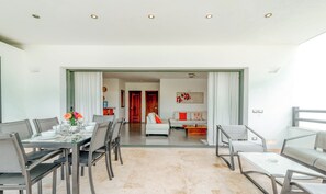 A modern dining area with a glass table and chairs, a sofa in the background, and a view into a living room.