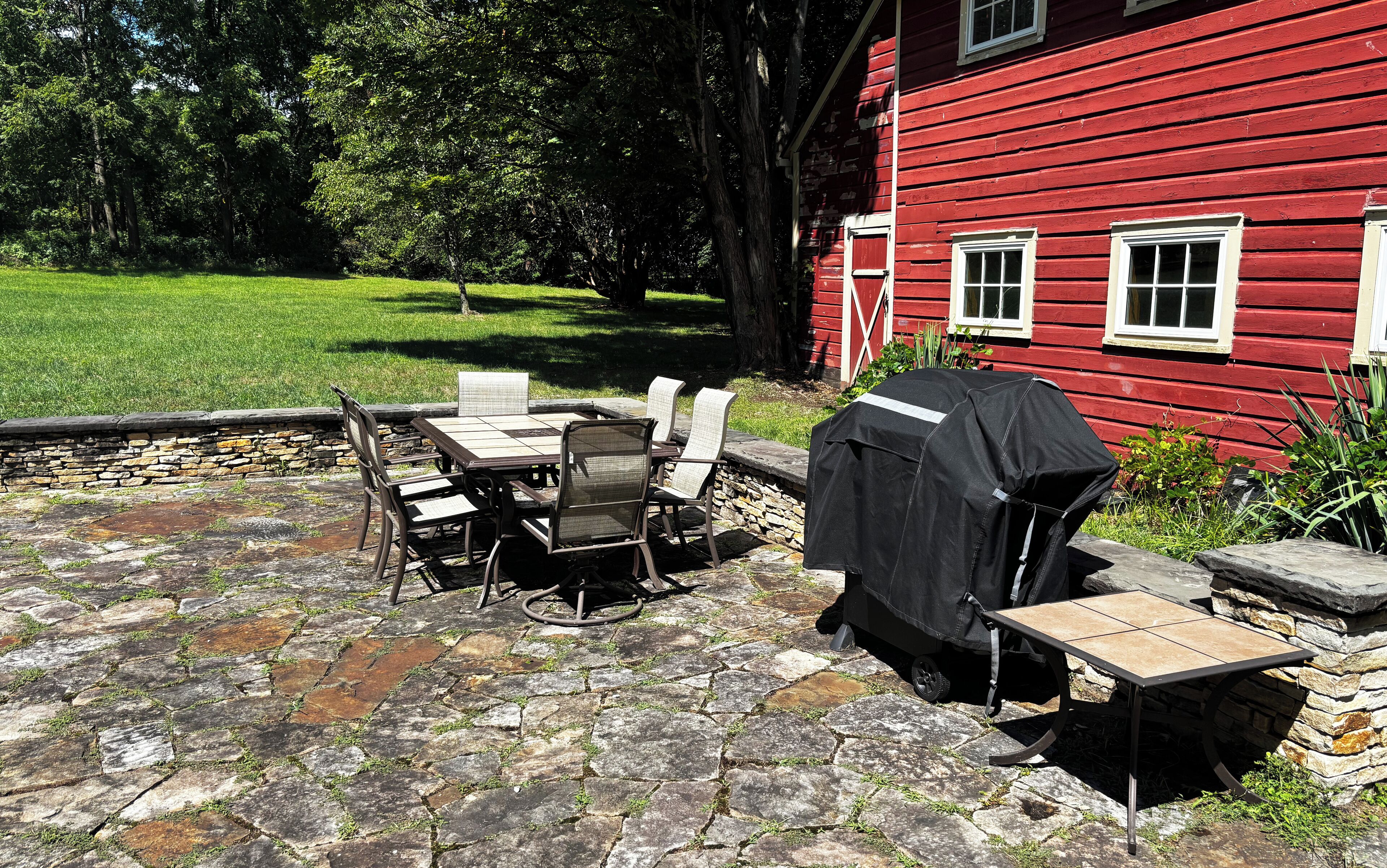 A stone patio with a table and chairs, a barbecue grill, and a red building in the background.