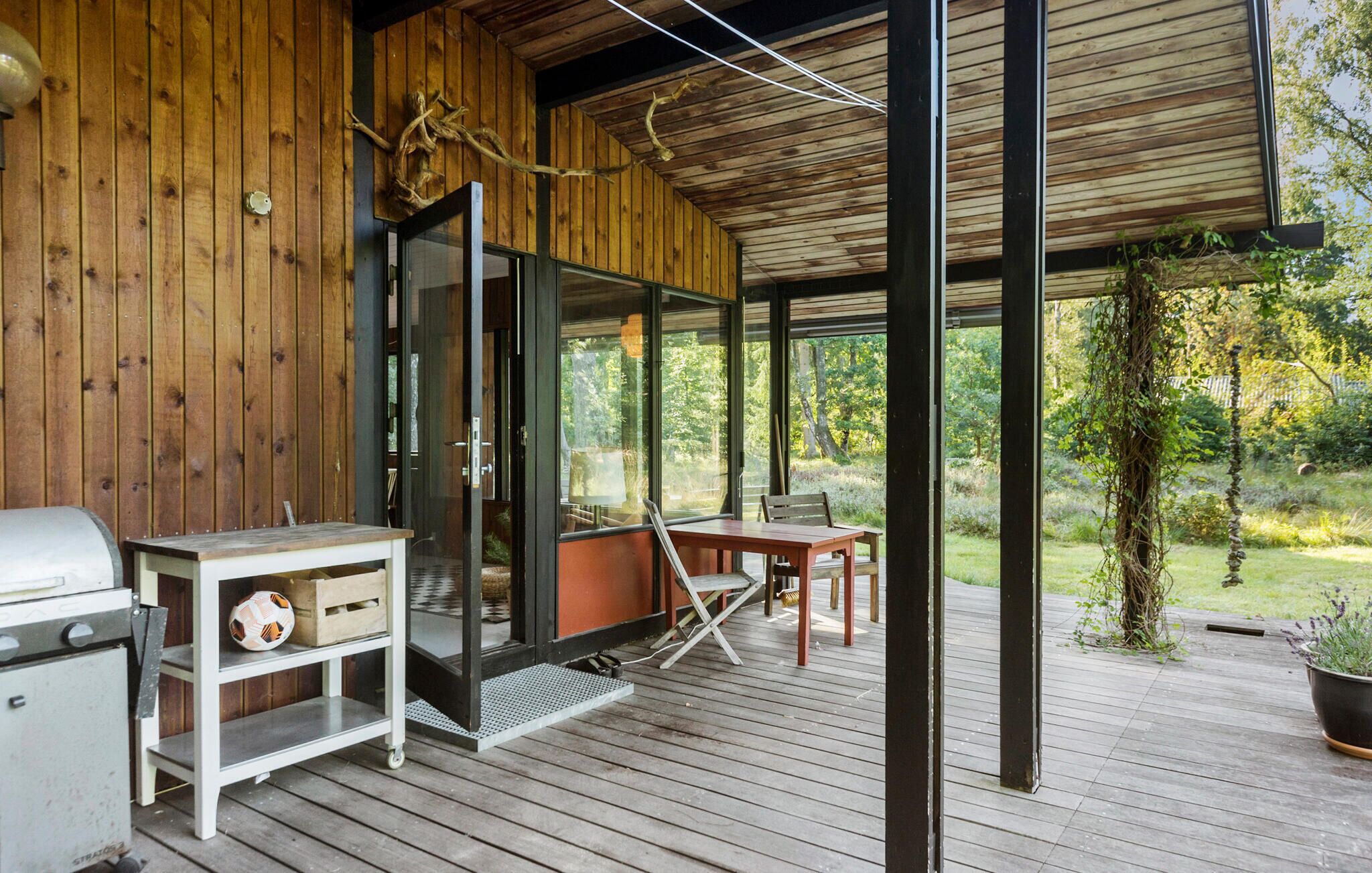 Eine Holzterrasse mit Glastür, die zu einem überdachten Außenbereich führt, einem Grill und einem Tisch mit Stühlen.