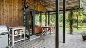 Een houten terras met een glazen deur naar een overdekt buitenverblijf, een barbecue en een tafel met stoelen.