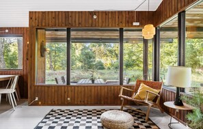 Une pièce aux murs lambrés, de grandes fenêtres donnant sur une forêt, un tapis à carreaux, un pouf en osier et un fauteuil en osier avec des coussins.