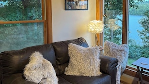 Una habitación acogedora con un sofá de cuero, ventanas amplias, un cuadro enmarcado y una mesa con una lámpara.