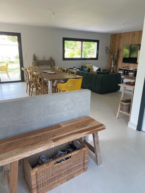 A modern living room with a wooden bench, dining table, and chairs. There is a large window providing natural light and a view of greenery outside.